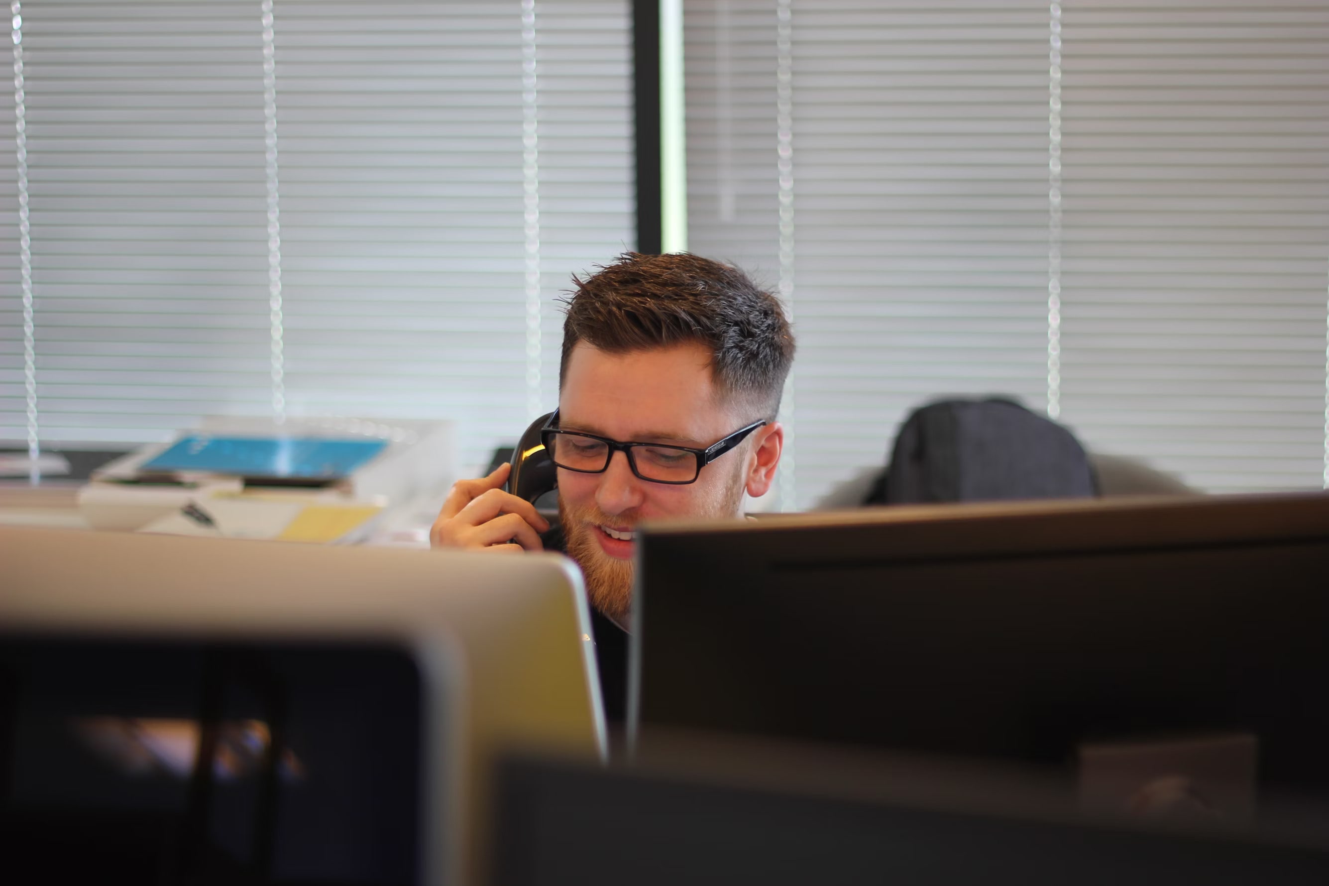 A man answering a telephone partially obscured by two computer monitors.